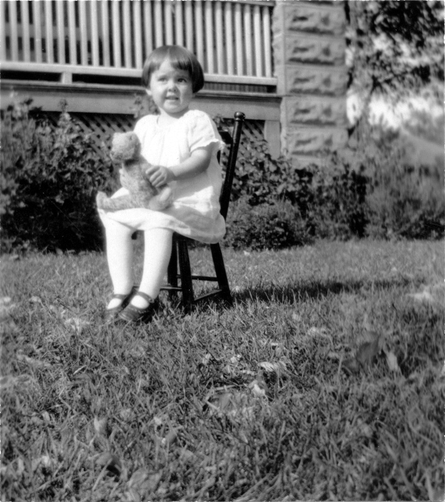 Black and white photograph of a young girl, seated on a chair outside, holding a teddy bear.
